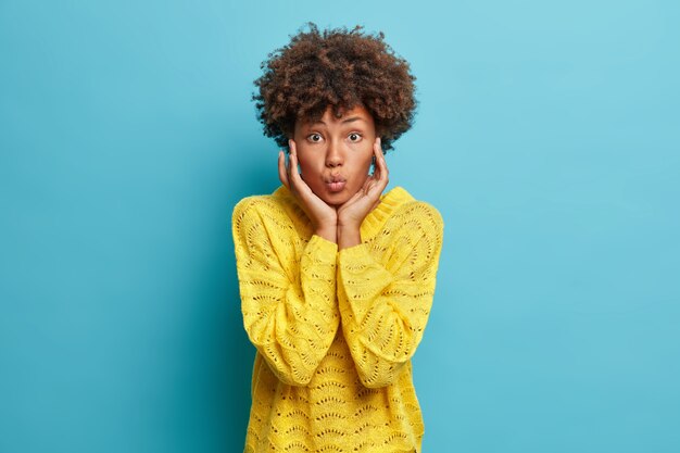 Coup de demi-longueur de la belle jeune femme afro-américaine touche le visage doucement garde les lèvres pliées a un regard tendre porte un pull en tricot jaune décontracté isolé sur un mur bleu veut embrasser quelqu'un