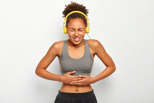 Coup de demi-longueur de femme insatisfaite aux cheveux bouclés, écoute de la musique avec des écouteurs, souffre de crampes douloureuses dans le ventre, isolé sur fond blanc