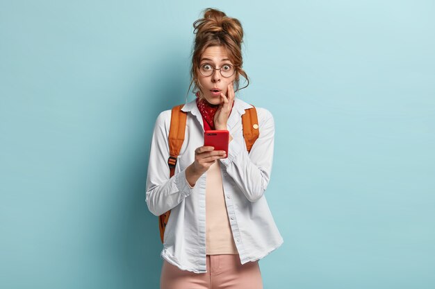 Coup de demi-longueur de jeune femme impressionnée avec expression effrayée, détient un téléphone portable moderne, étant voyageur auto-stoppeur