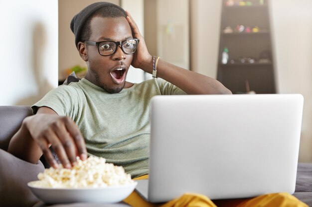 Coup franc d'un drôle de jeune homme à la peau sombre en lunettes et chapeau regardant match de football en ligne, en utilisant un ordinateur portable et en mangeant du pop-corn, assis sur un confortable canapé gris à la maison, touchant le visage sous le choc