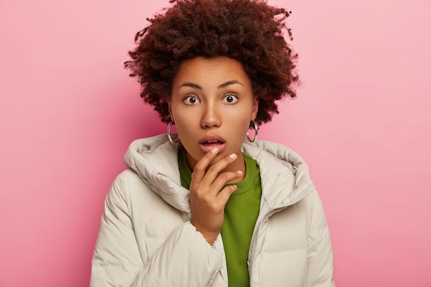 Photo gratuite coup franc d'une femme surprise en tenue d'hiver met la main sur la mâchoire tombée, regarde avec étonnement et isolé sur fond rose.