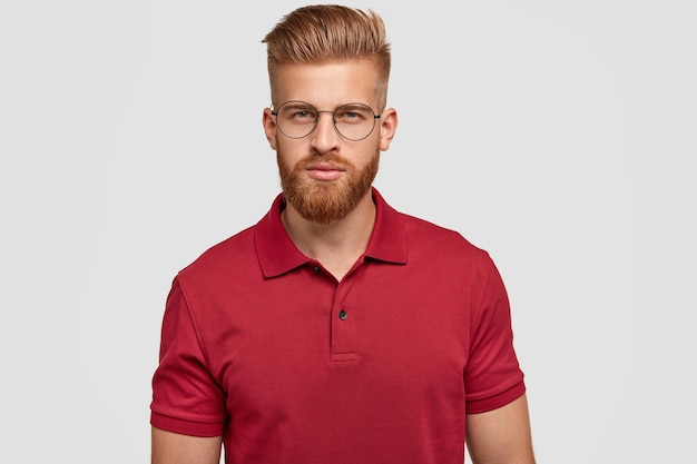 Coup franc d'un homme de race blanche sérieux et confiant avec des cheveux et une barbe foxy, habillé avec désinvolture, regarde directement avec une expression mystérieuse, porte des lunettes, isolé sur un mur blanc