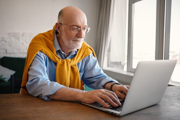Coup franc d'un homme de soixante ans, élégant et élégant, à la mode, avec une barbe grise et une tête chauve ayant un regard concentré, utilisant un ordinateur portable générique wifi, tapant rapidement. Concept de personnes, d'âge et de gadgets