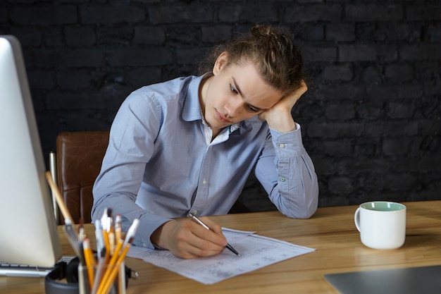 Coup franc d'un jeune architecte européen élégant travaillant au bureau, vérifiant les dessins à l'aide d'un stylo, ayant un regard triste et sérieux, se sentant fatigué et somnolent, articles de papeterie, tasse et ordinateur sur un bureau en bois