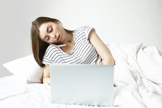 Coup franc d'une jeune femme d'affaires européenne sérieuse utilisant un ordinateur portable générique et un mobile tout en travaillant à distance, allongée dans son lit, parlant au téléphone, ayant concentré l'expression du visage