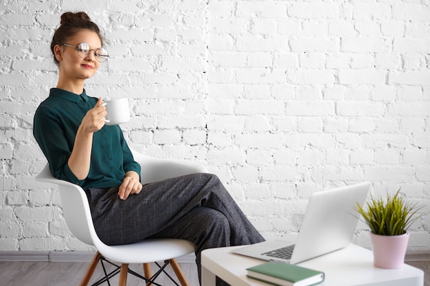 Coup franc de jeune pigiste à la mode portant des lunettes rondes et un chignon appréciant le café ou le thé à l'espace de coworking, assis sur une chaise devant un ordinateur portable ouvert, souriant