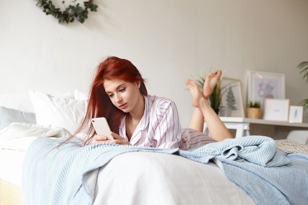 Photo gratuite coup franc d'une jolie jeune femme aux cheveux roux de 18 ans en pyjama rayé allongée sur le ventre sur le ventre sur le lit défait, les pieds en l'air, publiant des photos via les réseaux sociaux et vérifiant le fil d'actualité