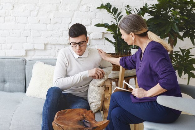 Coup franc d'une psychothérapeute professionnelle habillée avec désinvolture dans la cinquantaine, touchant son jeune patient de sexe masculin par l'épaule pendant une séance de conseil, exprimant sa sympathie et son soutien