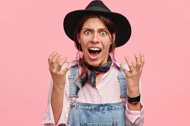 Coup horizontal de beaux gestes de travailleuse agricole agacée, a irrité l'expression du visage, mécontent du travail, porte un chapeau noir et une salopette en denim isolé sur un mur rose