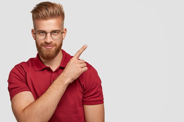 Photo gratuite coup de jeune homme séduisant au gingembre avec une barbe épaisse, pointe dans le coin supérieur droit, a une expression faciale confiante, porte un t-shirt rouge, isolé sur un mur blanc avec espace de copie