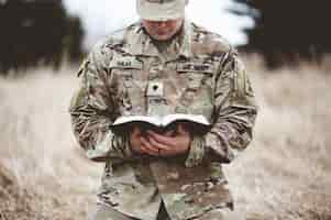 Photo gratuite coup de mise au point peu profonde d'un jeune soldat à genoux sur une herbe sèche en lisant la bible