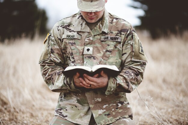 Coup de mise au point peu profonde d'un jeune soldat à genoux sur une herbe sèche en lisant la Bible