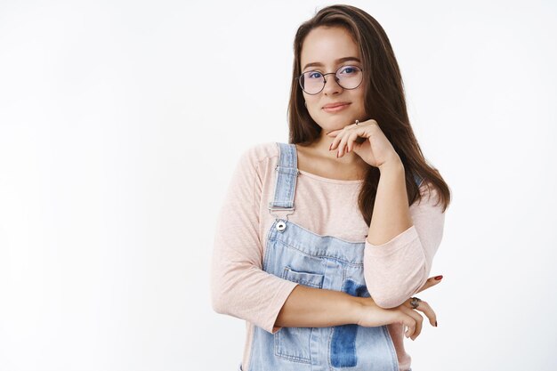 Coup de taille d'une artiste féminine intelligente et créative dans des lunettes et une salopette tenant la main sur la mâchoire et souriante ravie