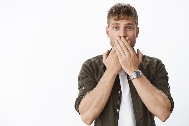 Coup de taille d'un beau mec blond choqué et sans voix entendant des nouvelles étonnantes indicibles couvrant la bouche avec les deux paumes et les yeux écarquillés