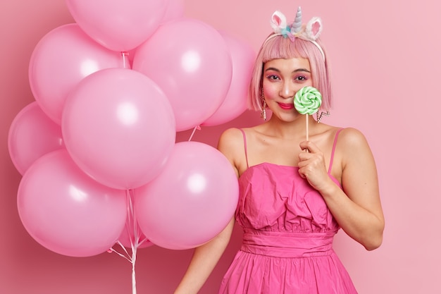 Coup de taille d'une femme asiatique assez heureuse tient des sourires de sucette douce a agréablement maquillage lumineux et la robe tient des ballons gonflés se prépare pour la célébration ou la fête isolée sur fond rose.