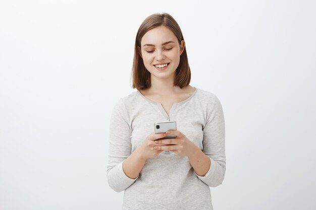 Coup de taille d'une femme brune européenne ordinaire moderne en tenue décontractée tenant un smartphone souriant à l'écran de l'appareil, envoyer des SMS ou modifier une photo pour publier en ligne posant sur un mur gris