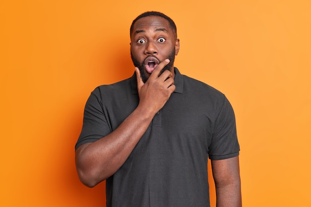 Photo gratuite coup de taille d'un homme choqué effrayé tient le menton et regarde les yeux écarquillés à l'avant vêtu d'un t-shirt noir surpris par quelque chose qui pose contre un mur orange vif