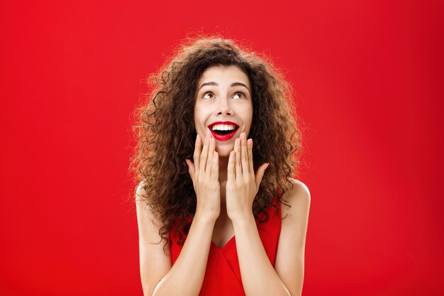 Coup de taille d'une jeune petite amie rêveuse amusée et amusée en robe élégante tenant les paumes sur le menton et ouvrant la bouche avec un large sourire semblant ravie comme si elle voyait un miracle sur un mur rouge.