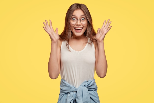 Coup de taille d'une jolie femme aux cheveux noirs avec une expression positive, écarte les mains en recevant la surprise de son petit ami, se sent heureux, porte des lunettes transparentes, un gilet décontracté, isolé sur un mur jaune