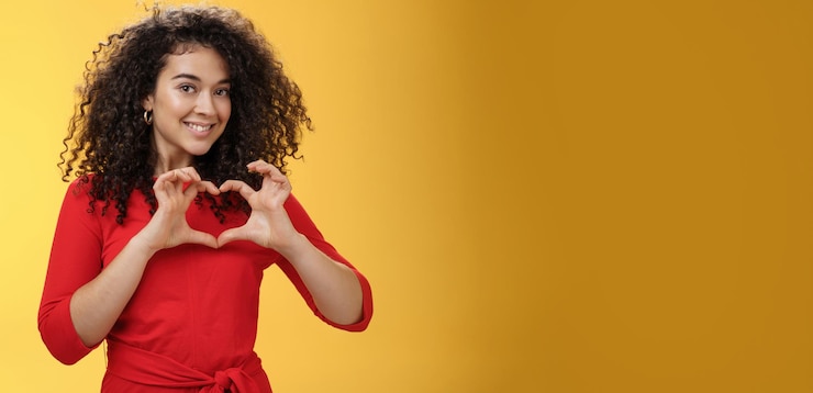 Photo gratuit coup de taille de jolie petite amie romantique et mignonne aux cheveux bouclés en robe rouge montrant le signe du coeur