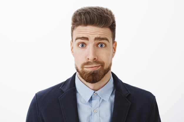 Coup de taille d'un petit ami brunet mignon stupide et sombre avec des yeux bleus et une barbe souriant debout en costume élégant se sentant désolé de s'excuser d'être stupide posant contre un mur gris