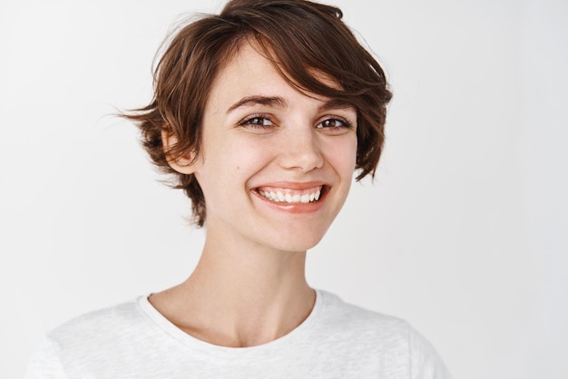 Coup de tête d'une jolie fille souriante avec une peau lisse et éclatante qui a l'air heureuse et pleine d'espoir debout sur fond blanc