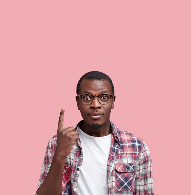 Coup vertical d'un beau mâle afro-américain confus avec une peau sombre et saine, porte une chemise et des lunettes, pose seul à l'espace de copie vierge, fait de la publicité pour quelque chose.