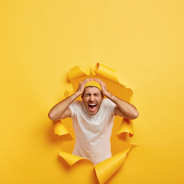 Coup vertical d'un gars émotionnel touche la tête avec les deux mains, porte un t-shirt blanc décontracté, un chapeau jaune élégant
