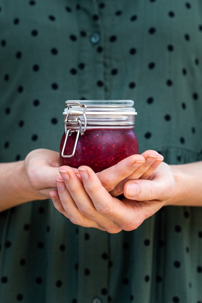 Coup vertical de mains féminines tenant une confiture de framboises crues vegan maison dans un bocal en verre