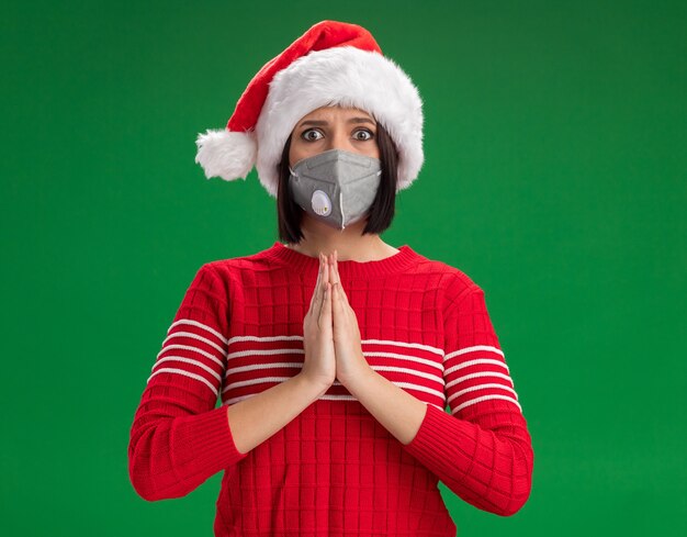 Coupable jeune fille portant bonnet de Noel et masque de protection regardant la caméra en gardant les mains en geste de prier isolé sur fond vert