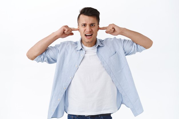 Coupez le volume. Portrait d'un bel homme agacé et dérangé qui a l'air irrité, ferme les oreilles avec les doigts d'un son gênant ennuyeux, de la musique forte ou des voisins, mur blanc