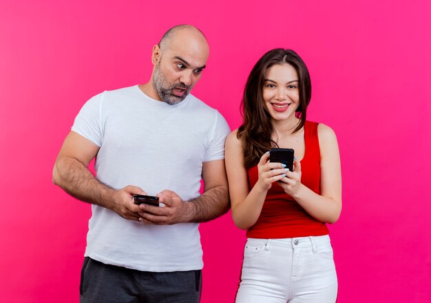 Couple adulte à la fois tenant des téléphones mobiles homme surpris regardant le téléphone de la femme et femme souriante à la recherche