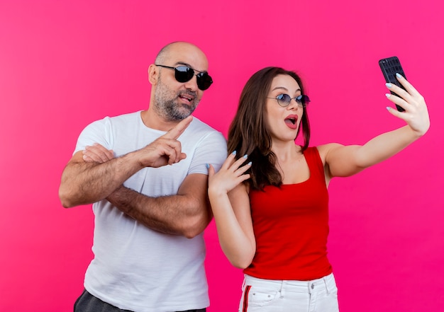 Couple d'adultes impressionnés portant des lunettes de soleil homme faisant signe de paix femme gardant la main dans l'air en prenant selfie