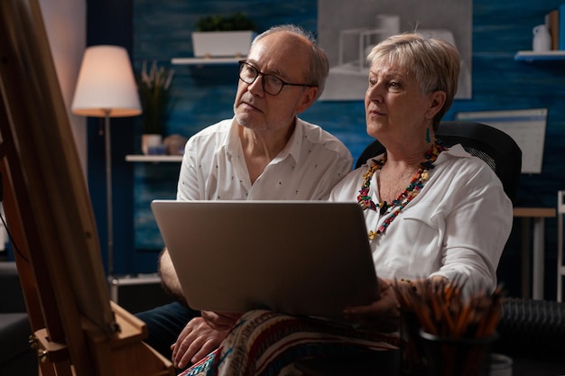 Couple âgé assis devant un chevalet tenant un ordinateur portable regardant des tutoriels en ligne pour améliorer la technique de dessin. Homme et femme à la retraite à la recherche d'inspiration sur les réseaux sociaux à l'aide d'un ordinateur portable.