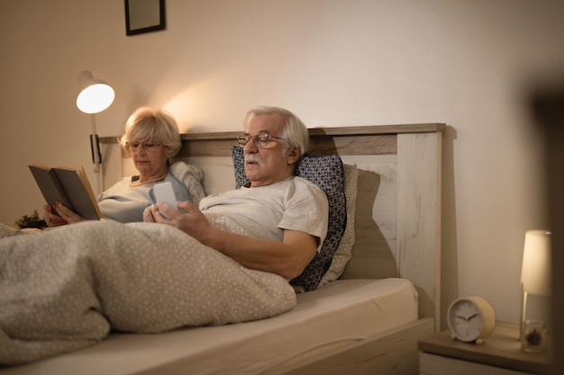 Couple d'âge mûr se détendre au lit le soir L'accent est mis sur l'homme utilisant un téléphone intelligent pendant que sa femme lit un livre