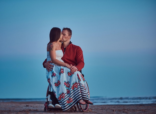 Photo gratuite un couple d'amoureux, une femme assise sur les genoux de son mari, s'embrassant sur une plage sur fond d'aube lumineuse.