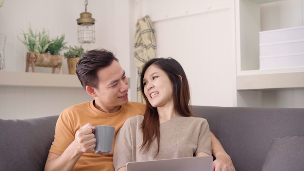 Couple asiatique, utilisation, ordinateur portable, et, boire, chaud, tasse café, dans, salon, à, maison
