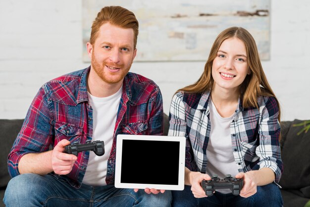 Couple assis sur le canapé tenant la manette de jeu dans les mains montrant une tablette numérique vierge