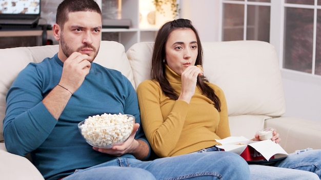 Couple assis sur un canapé en train de manger du poulet frit et du pop-corn en regardant la télévision. Couple effrayé après un moment effrayant dans le film.