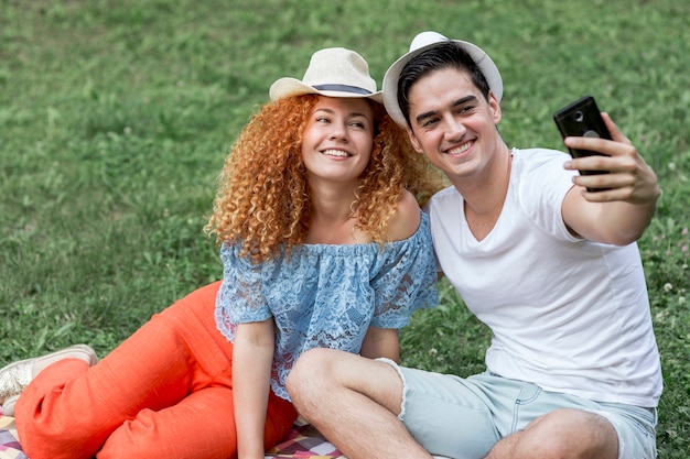 Photo gratuite couple assis sur une couverture de pique-nique et prenant un selfie