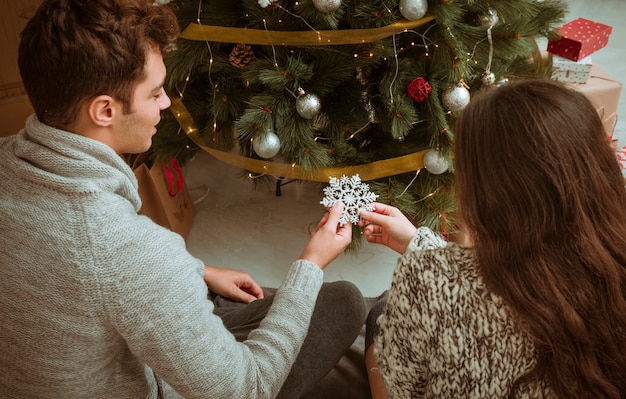 Couple assis sous un sapin de Noël tenant un flocon de neige ornemental