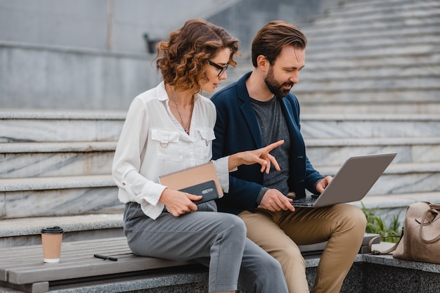 Couple attrayant d'homme et de femme parlant assis dans les escaliers du centre-ville urbain, travaillant ensemble sur un ordinateur portable