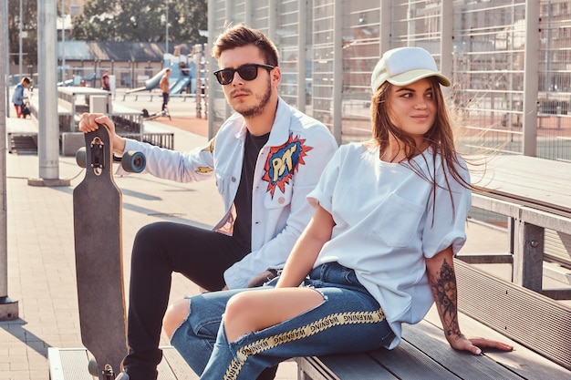 Photo gratuite couple attrayant de jeunes hipsters habillés à la mode avec des planches à roulettes assis sur un banc au complexe sportif de la ville le jour ensoleillé, avec des tons chauds.