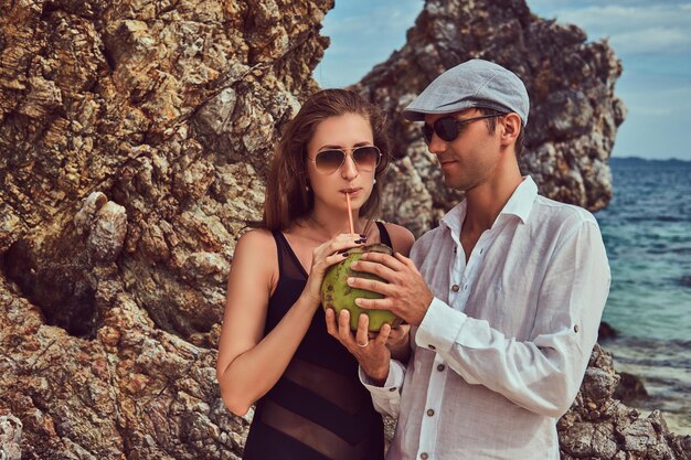 Un couple attrayant, tenant une noix de coco avec un tube, debout sur une plage près de grosses pierres de récif, profite de vacances sur une belle île.