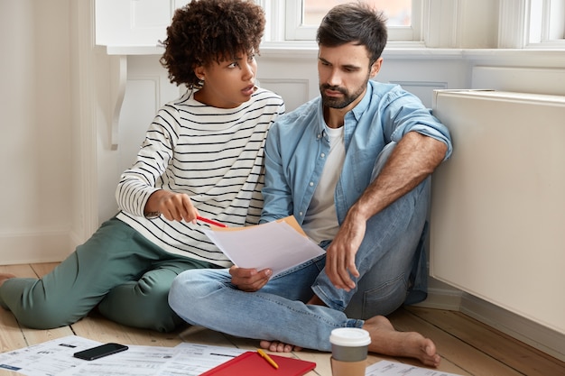 Un couple calcule ses factures à la maison, regarde désespérément ses papiers, a beaucoup d'impôts impayés, vérifie la documentation, pose sur le sol, boit du café dans un gobelet jetable, pense à réparer un appartement