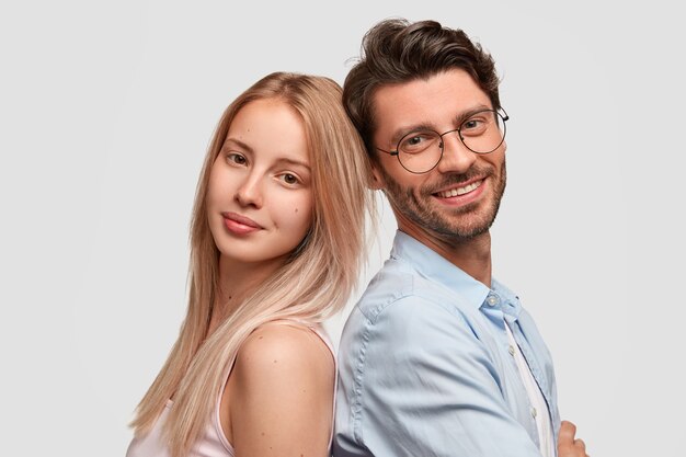 Un couple charmant se recule, étant de bonne humeur, isolé sur un mur blanc. Jolie femme blonde, homme gai mal rasé à lunettes étant des partenaires, se sentent soutenus, prêts à aider