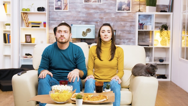 Couple déçu après que leur équipe de football préférée ait perdu le match. Chat allongé sur le canapé. Pop-corn, pizza et soda sur table basse.