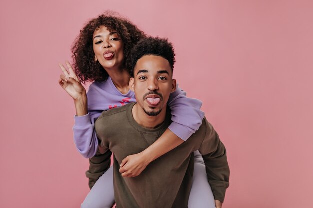 Couple drôle amoureux montrant des langues. Fille en tenue violette montrant un signe de paix sur un mur rose