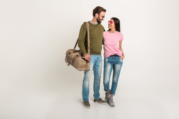 Couple élégant isolé, jolie femme souriante en t-shirt rose et homme en sweat-shirt tenant un sac de voyage, vêtu de jeans, portant des lunettes de soleil, s'amuser ensemble