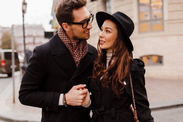Couple élégant à la mode en plein air amoureux embarrassant et se regardant avec adoration. Femme brune en bonnet de laine avec son petit ami en écharpe et manteau.